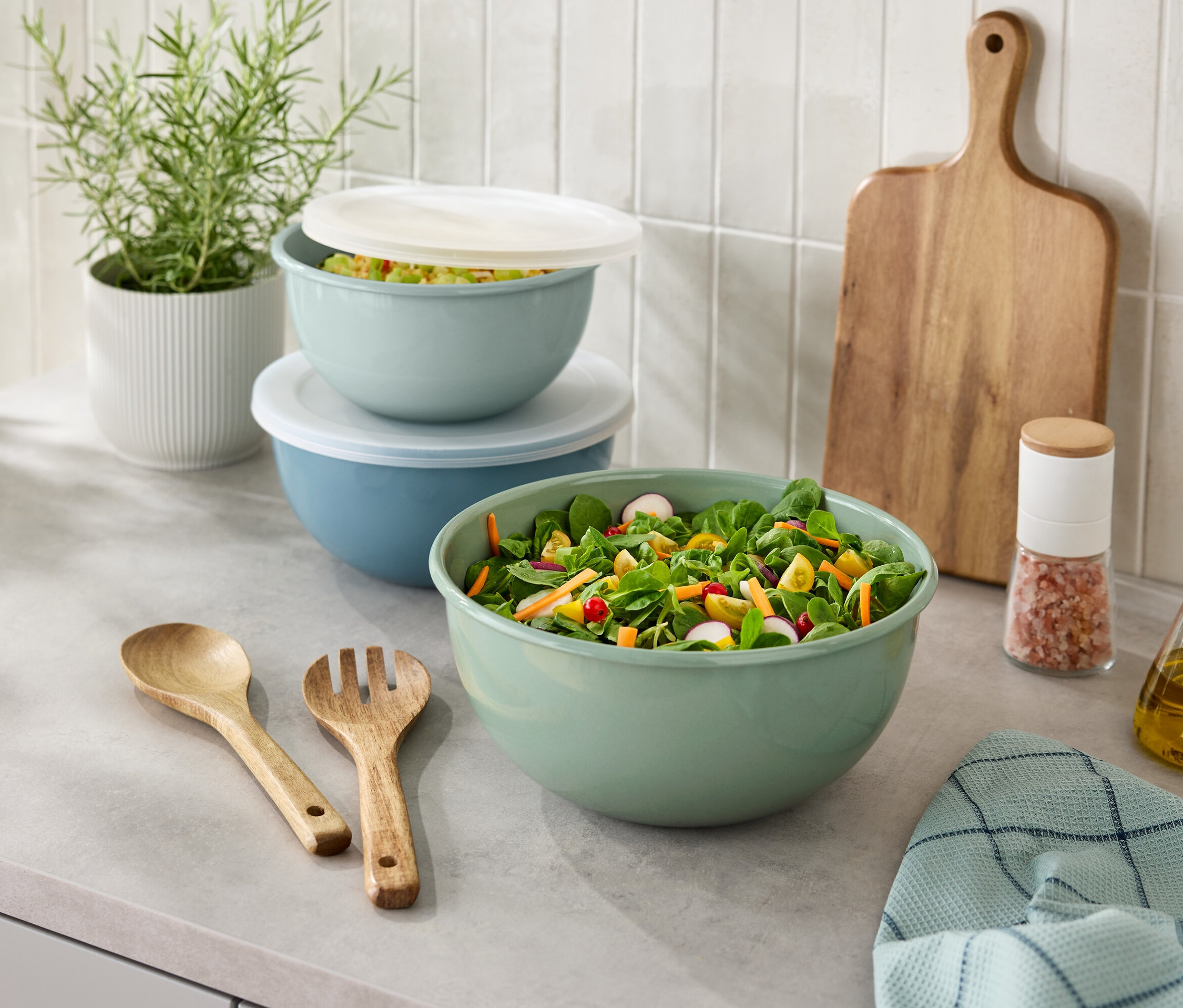 Tezgahta salata kasesi, tahta mutfak eşyaları, tuzluk, yağ şişesi, kesme tahtası ve saksıda bitki duruyor.