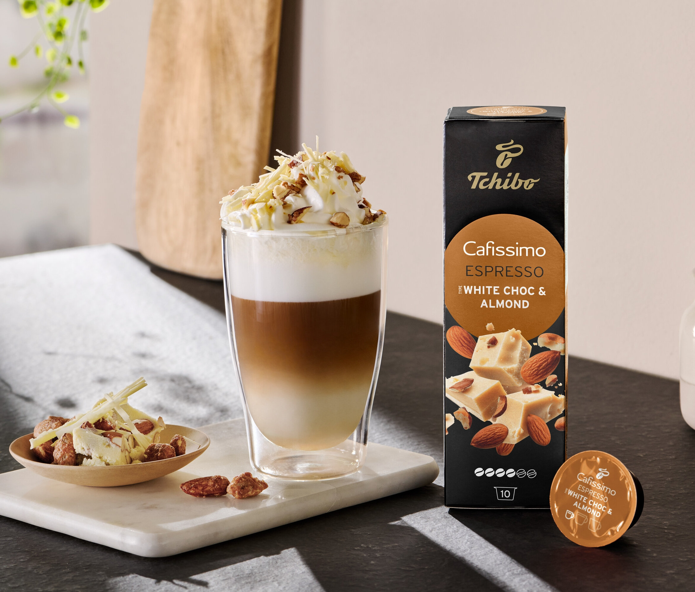 Katmanlı latte, krem şanti ve bir paket Cafissimo Flavoured Espresso - White Choc & Almond'un yanında serpilmiş halde.