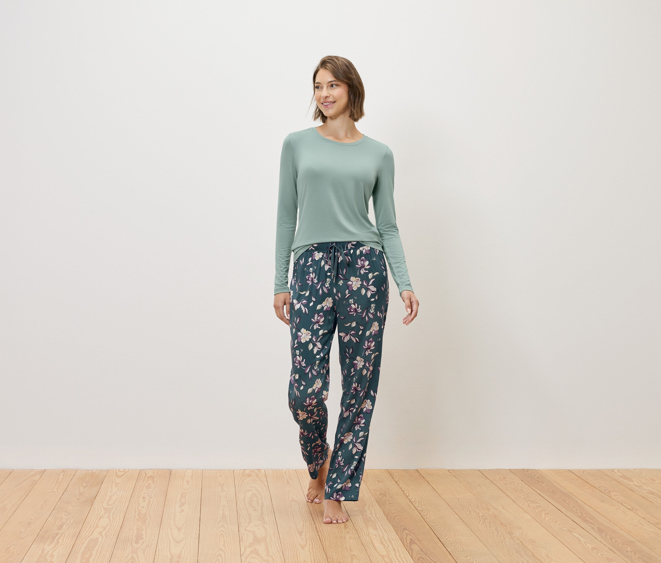 Bir kadın yalın ayak yürüyor, üzerinde nane yeşili bir kazak ve çiçek desenli lacivert bir pijama altı var.