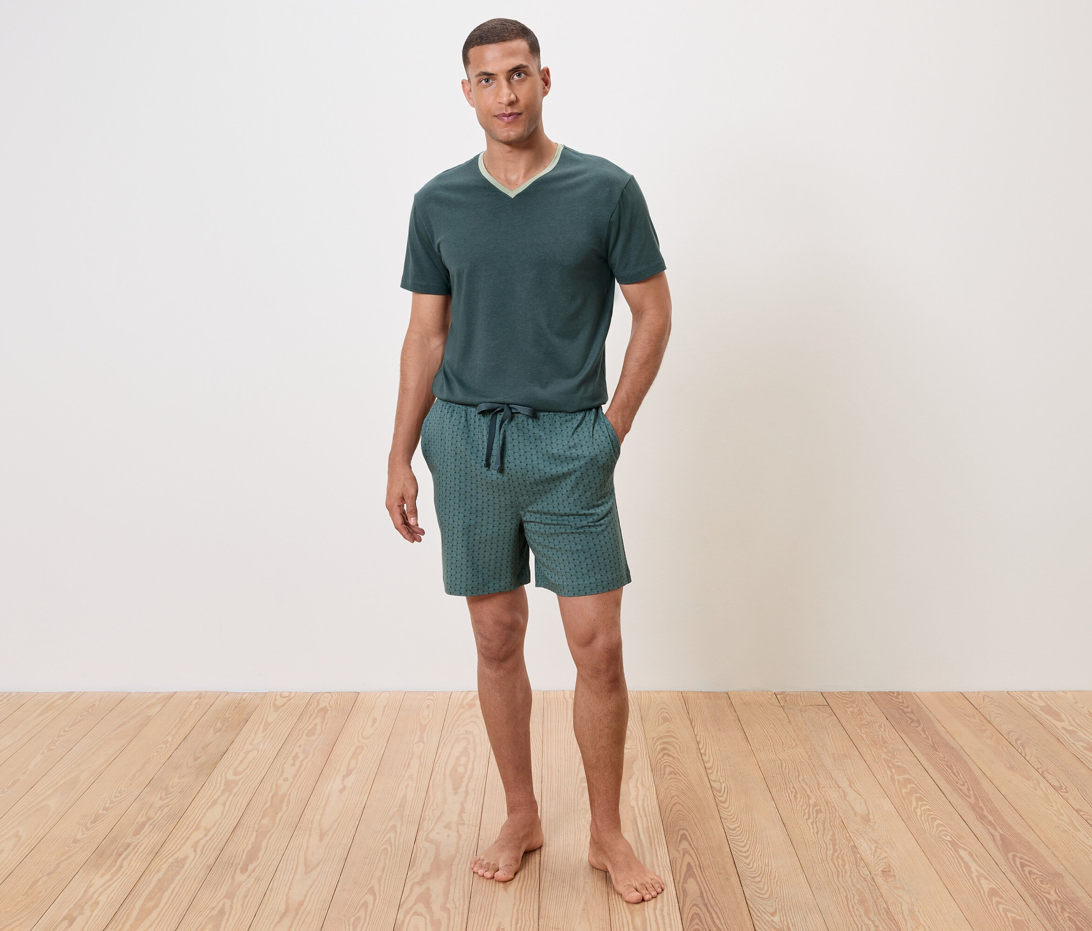 Adam yerde çıplak ayakla duruyor, kısa kollu yeşil bir pijama ve şort giyiyor.