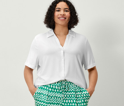 Bir kadın baskılı 3/4 jarse pantolon ve beyaz düğmeli bluzu ile ayakta duruyor, elleri cebinde.