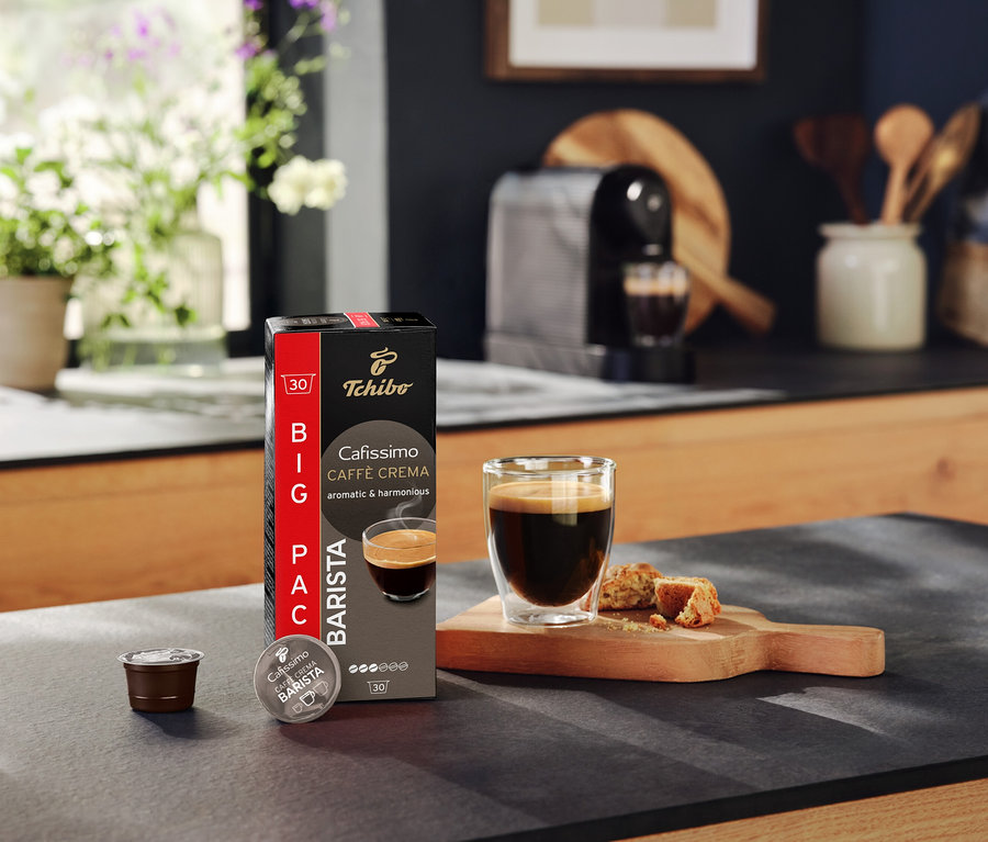 Mutfak tezgahında bir paket Tchibo Cafissimo Caffè Crema, bir kapsül, bir fincan kahve ve hamur işi duruyor.