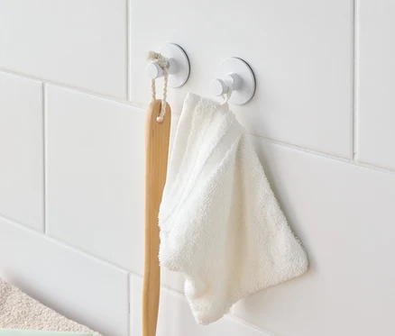Beyaz fayanslı duvarda beyaz kancalarda asılı bir banyo fırçası ve beyaz bir havlu.
