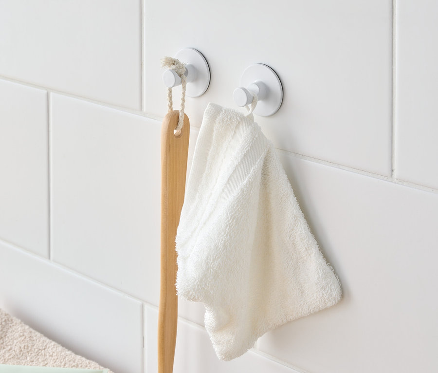 Beyaz fayanslı duvarda beyaz kancalarda asılı bir banyo fırçası ve beyaz bir havlu.