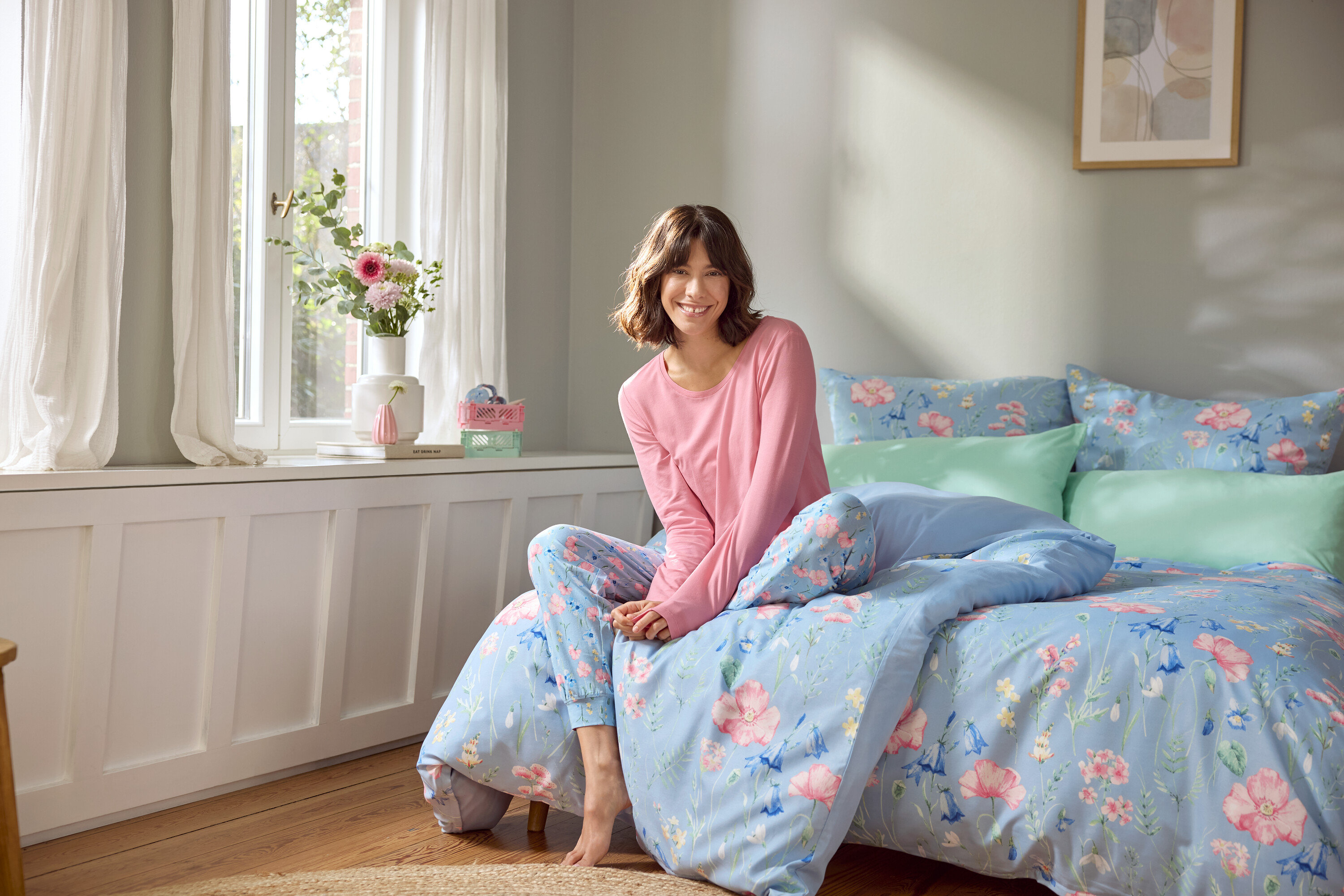 Kadın çiçek desenli açık mavi nevresimli bir yatakta oturuyor.