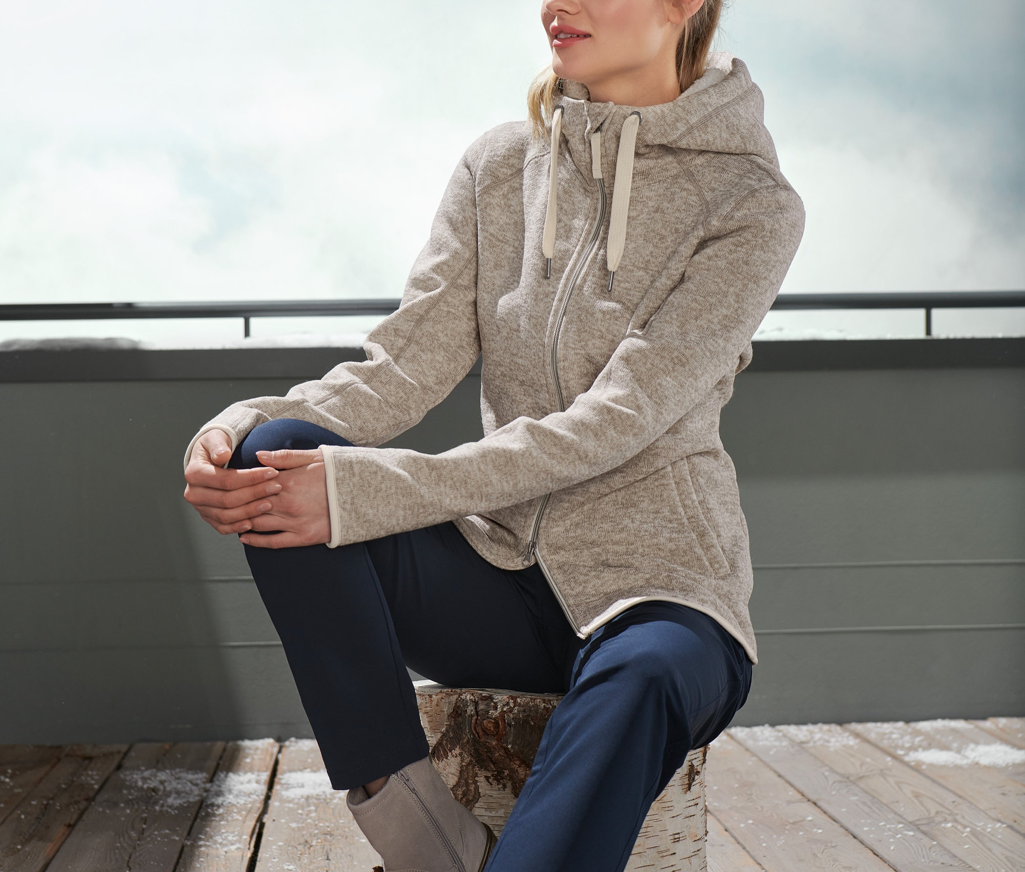 Kadın bir kütük üzerinde oturuyor ve bej rengi peluş örgü polar hırka giyiyor.