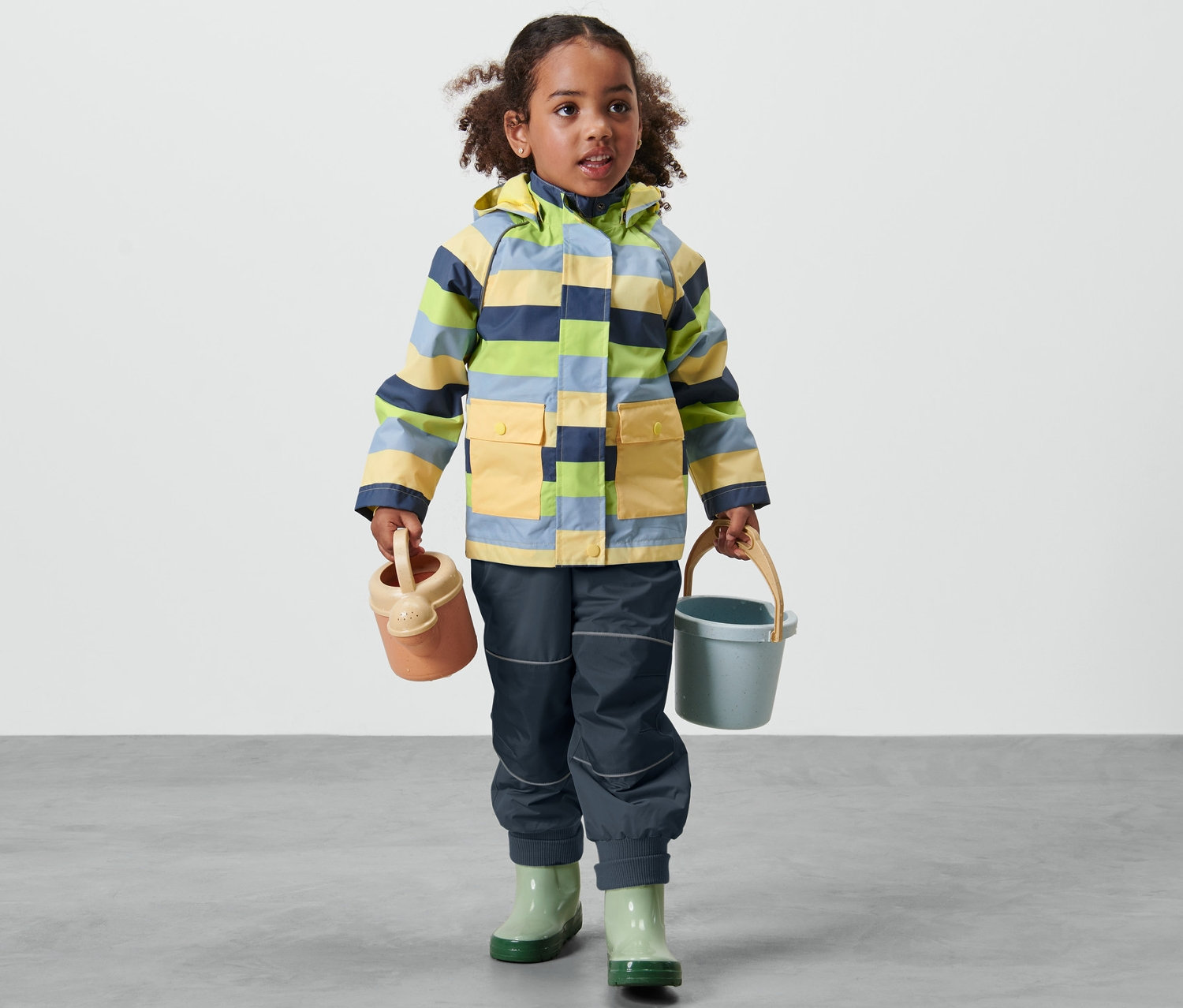 Çizgili çocuk yağmurluğu giyen kız çocuğu sulama kabı ve kova tutuyor.
