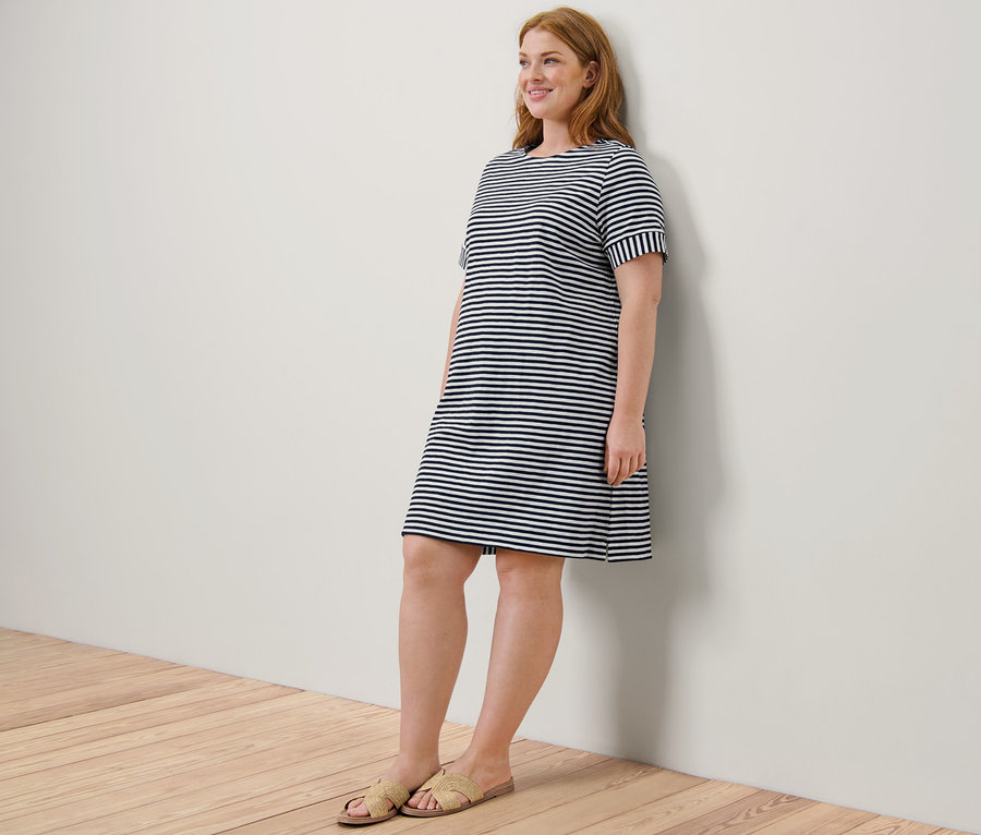 Kızıl saçlı bir kadın, kum rengi sandaletleri ve Çizgili Jersey Elbisesiyle bir duvara yaslanırken gülümsüyor.