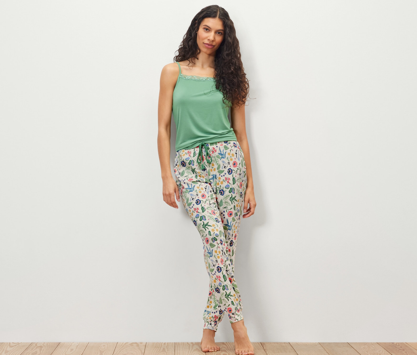 Beyaz bir duvarın önünde duran bir kadın, yeşil bir bluz ve pijama altı giyiyor.