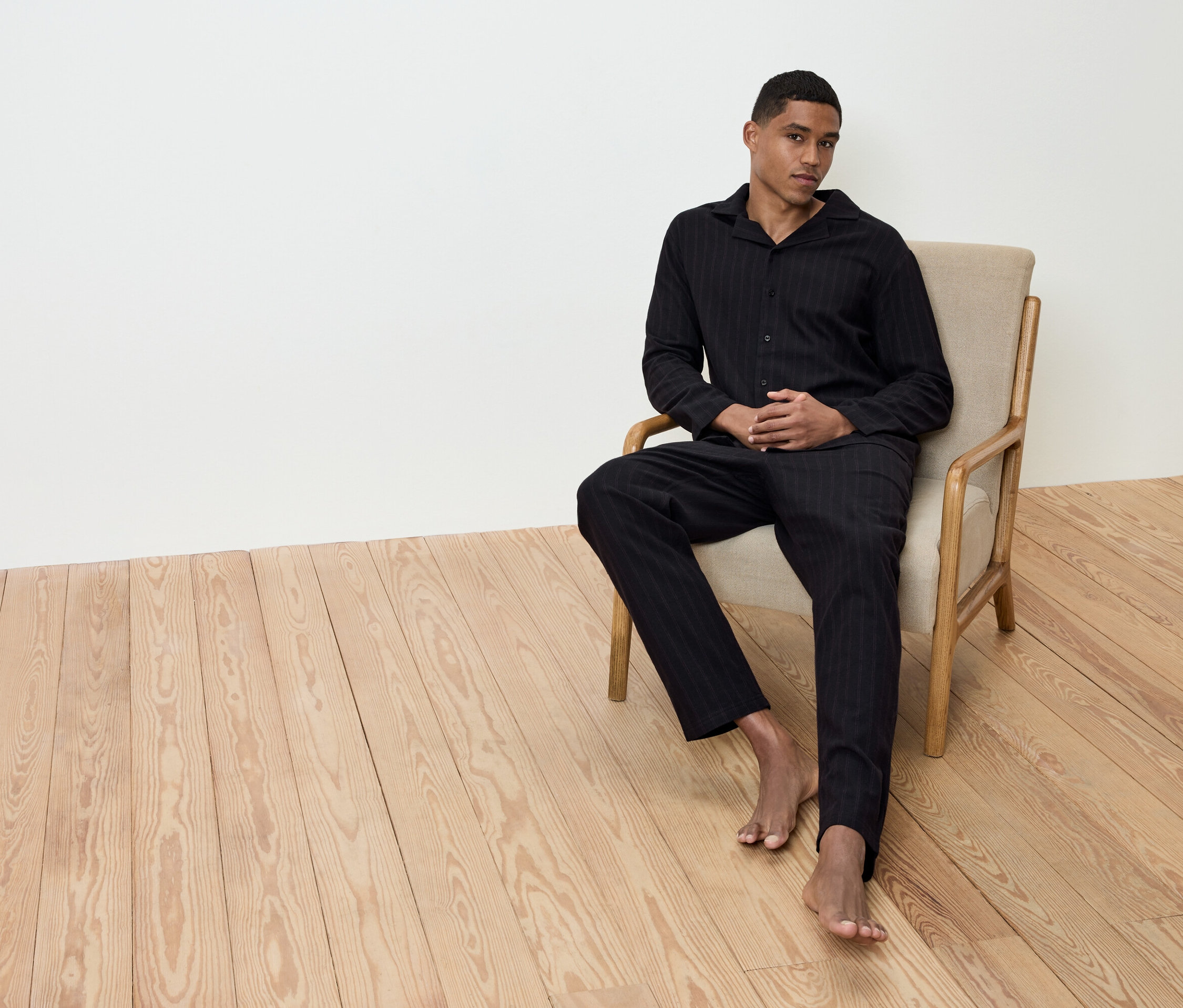 Adam yerde çıplak ayakla bej rengi bir koltukta oturuyor, üzerinde siyah çizgili bir pijama var.