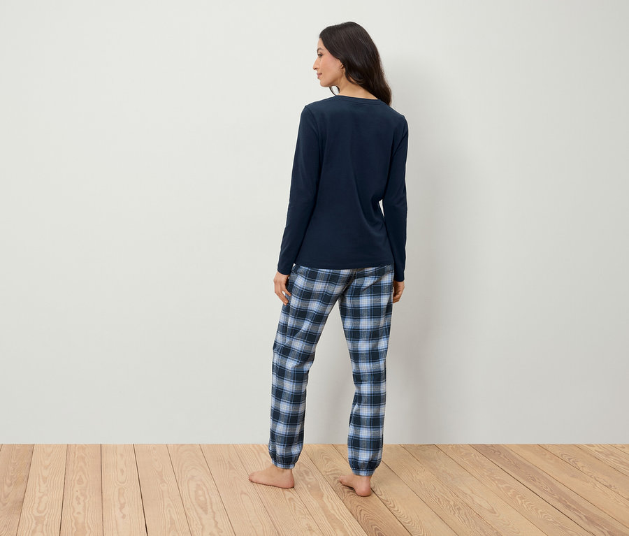 Kareli pantolonlu, lacivert pijamalı bir kadın, beyaz bir duvarın önünde ahşap bir zeminde kameraya arkası dönük duruyor.