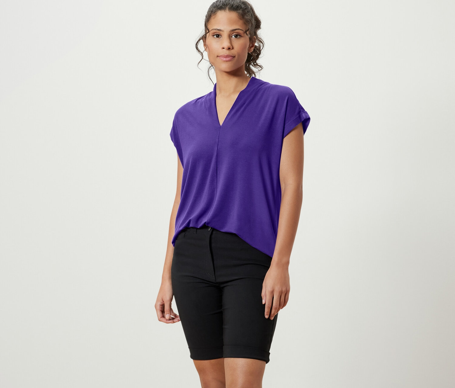 Bir kadın ayakta duruyor, üzerinde mor V yakalı bluz, siyah şort ve beyaz spor ayakkabılar var.