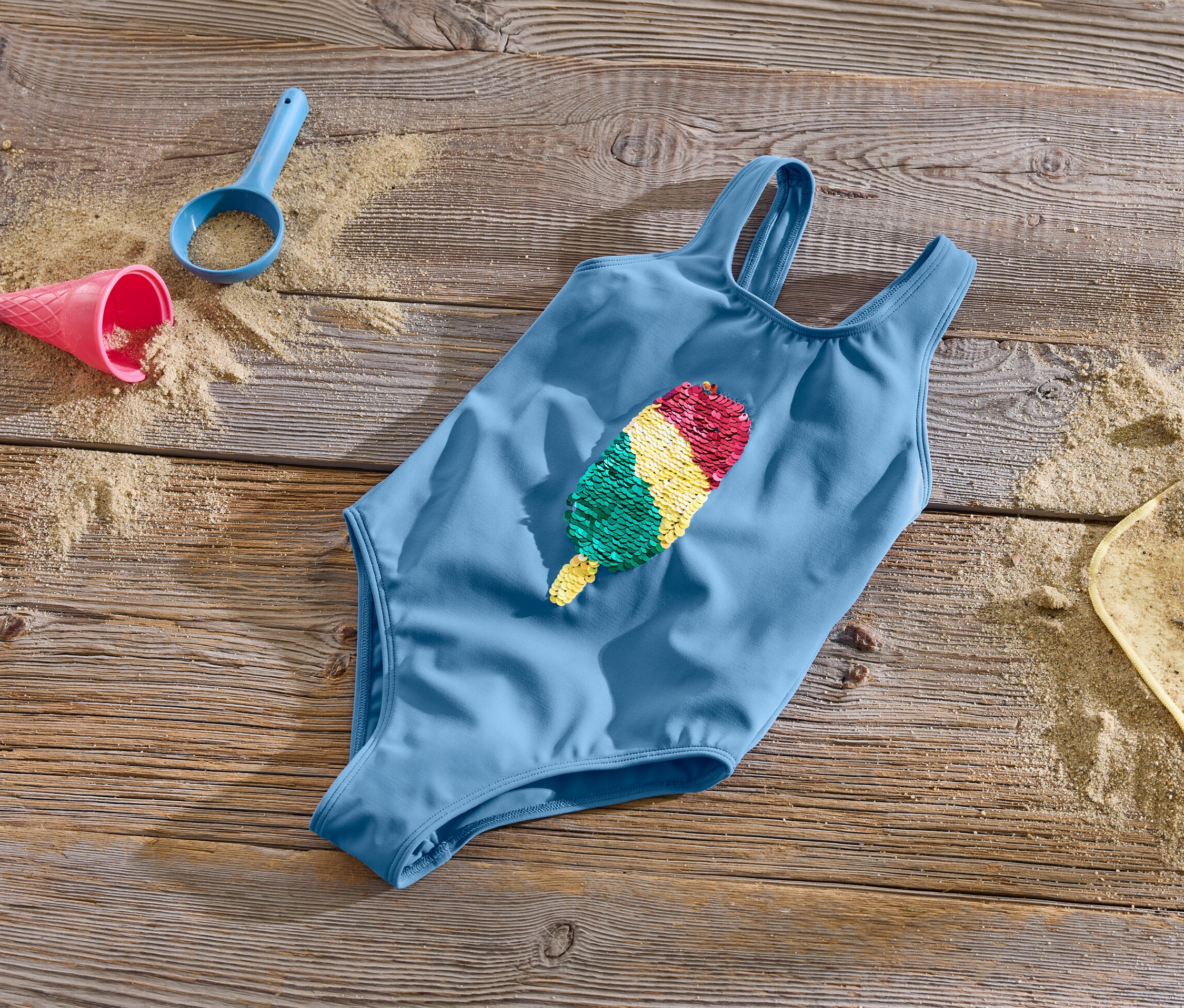 Mavi Çift Yönlü Payetli Çocuk Mayo, kum ve oyuncaklarla birlikte ahşap bir yüzeyde duruyor.