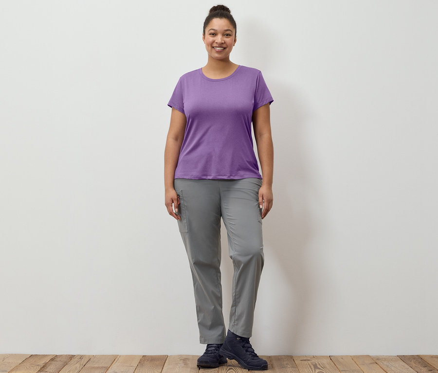 Bir kadın ayakta duruyor, gri, kendi cebine toplanabilir outdoor pantolonu ve fonksiyonel streç tişört giyiyor.