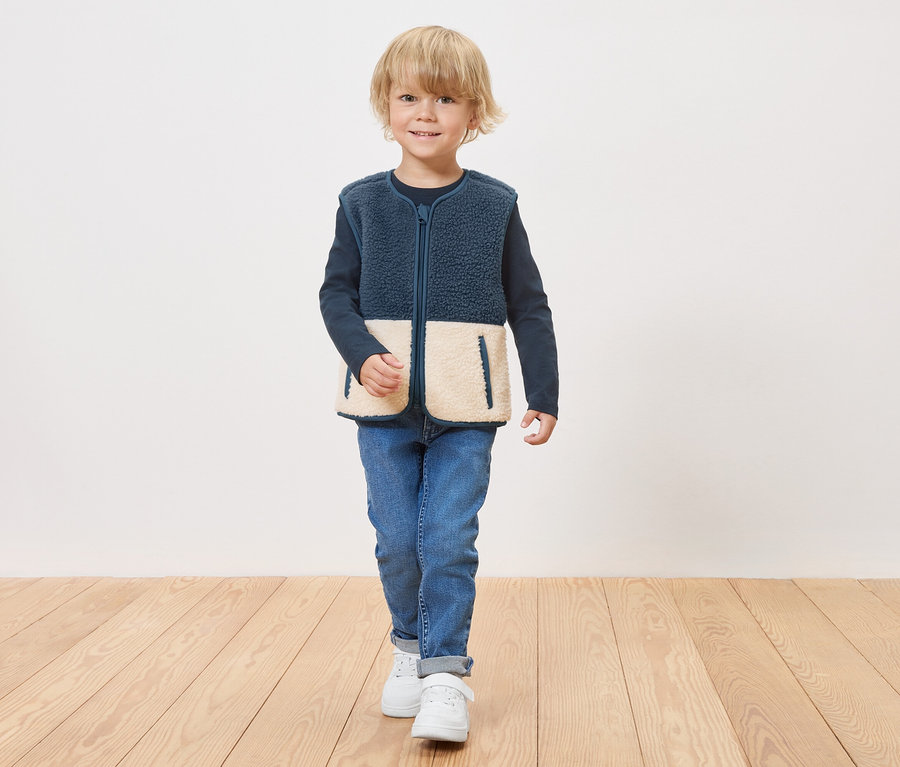 Lacivert renkli, krem alt panelli, fermuarlı polar yelek giyen bir çocuk ahşap zeminde duruyor.