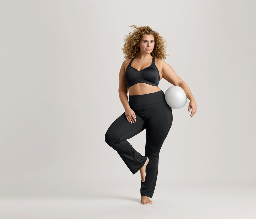 Kıvırcık saçlı bir kadın tek bacağı üzerinde dengede duruyor, beyaz bir top tutuyor, siyah bir sporcu sütyeni ve siyah tayt giyiyor.
