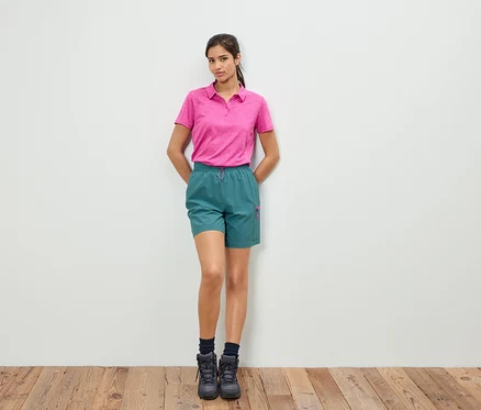 Bir kadın beyaz bir duvarın önünde duruyor, pembe bir polo tişört ve yeşil şort giyiyor.