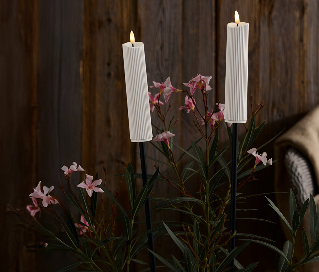 Koyu ahşap bir arka planın önünde pembe çiçekler ve yeşil yapraklar arasında siyah ayaklıklı iki adet beyaz, fitilli mum.