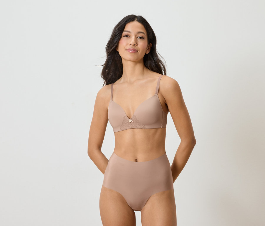 Kadın bej rengi bir sütyen ve külot giyiyor, her ikisi de beyaz bir arka planda sergileniyor.