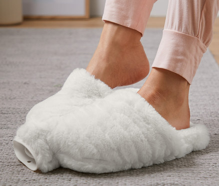 Gri halı üzerinde beyaz ayak ısıtıcı içinde ayaklar.