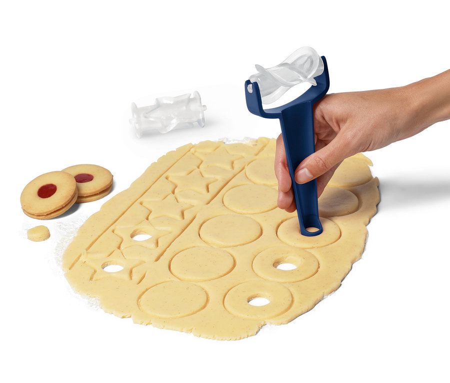 Bir el, hamur üzerinde bir kurabiye merdanesi kullanıyor. Yanında iki pişmiş kurabiye ve bir başlık duruyor.