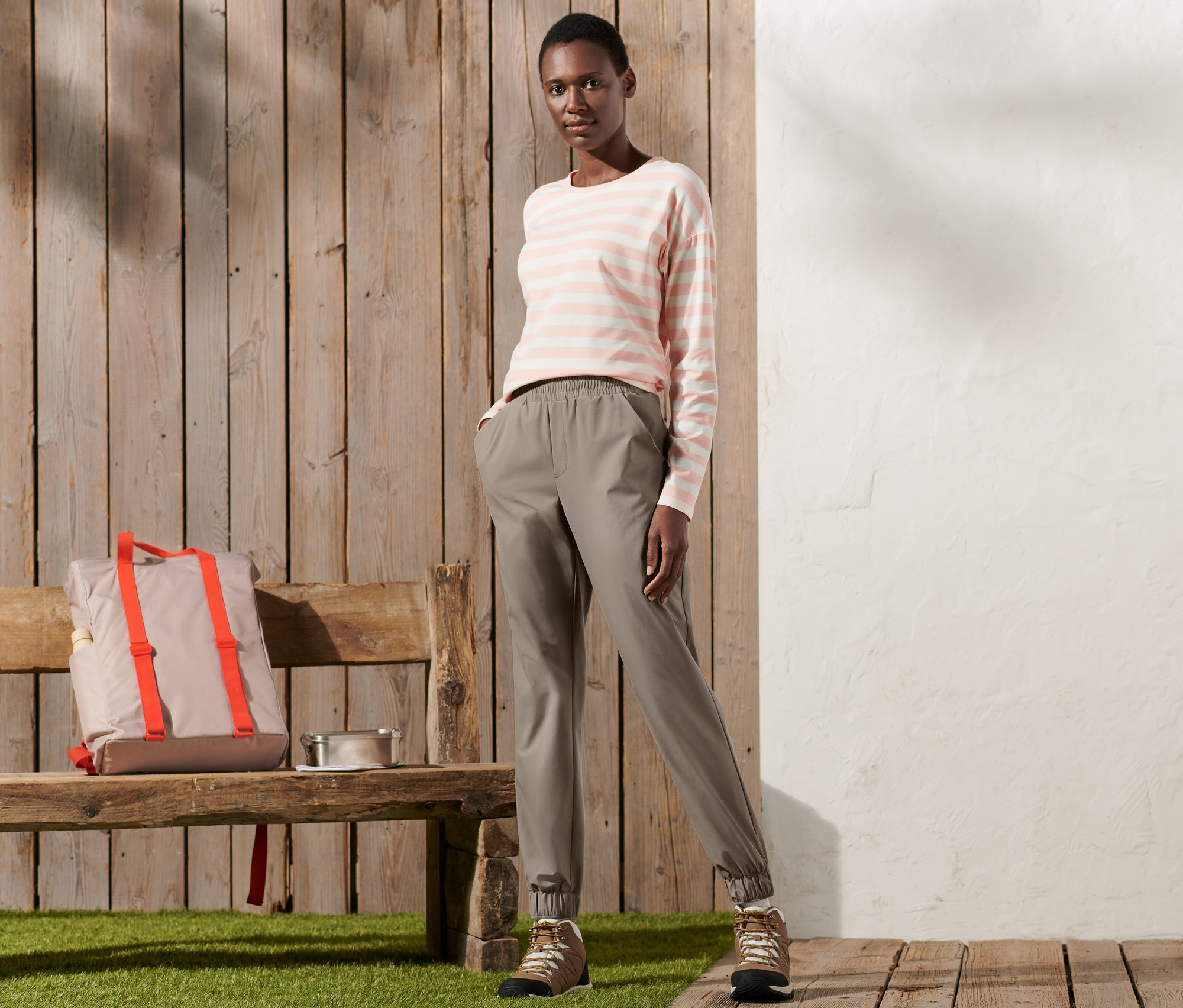 Bir kadın ahşap bir zeminde duruyor ve poz veriyor. Üzerinde bir fonksiyonel pantolon ve çizgili bir tişört var.