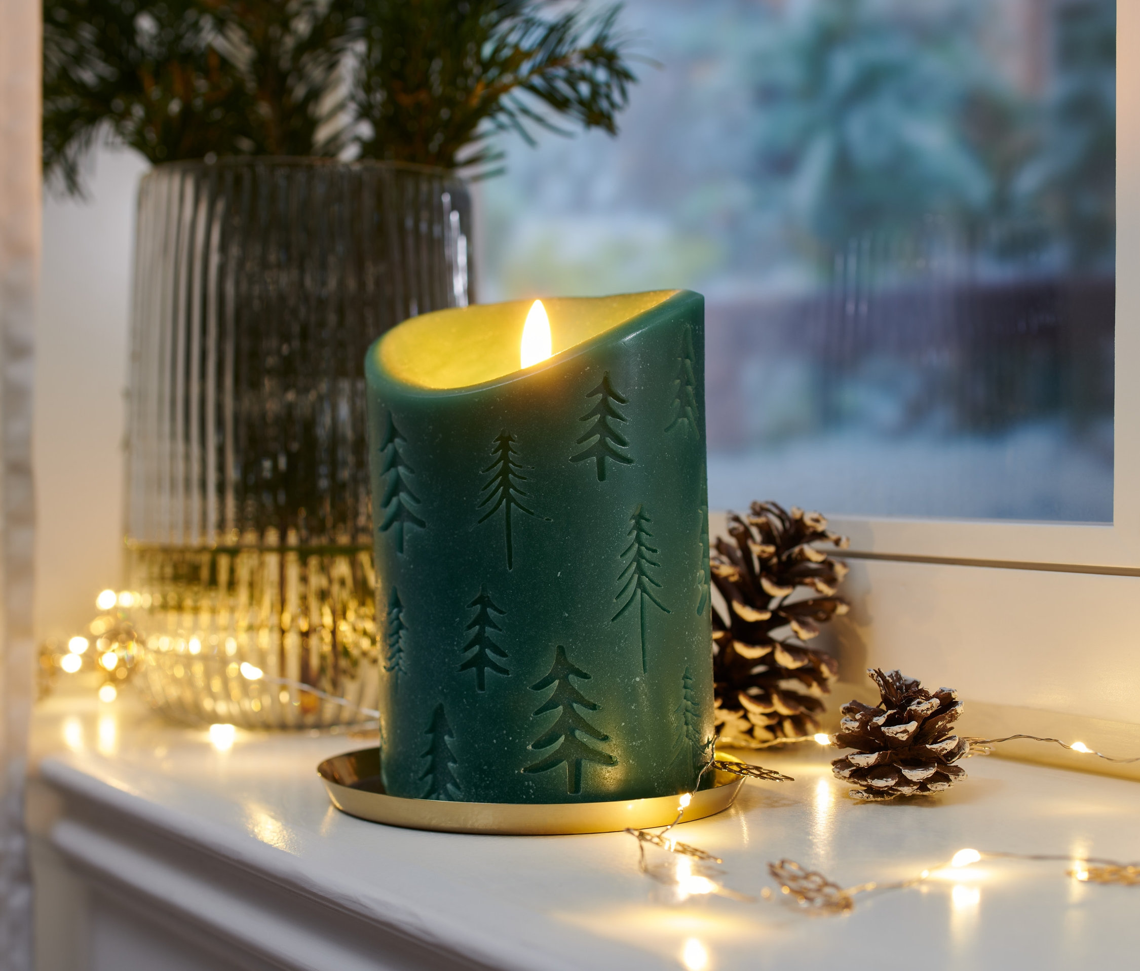 Çam ağacı desenli, altın renkli tabakta, kozalaklar ve ışıklarla birlikte duran yeşil renkli LED mum.