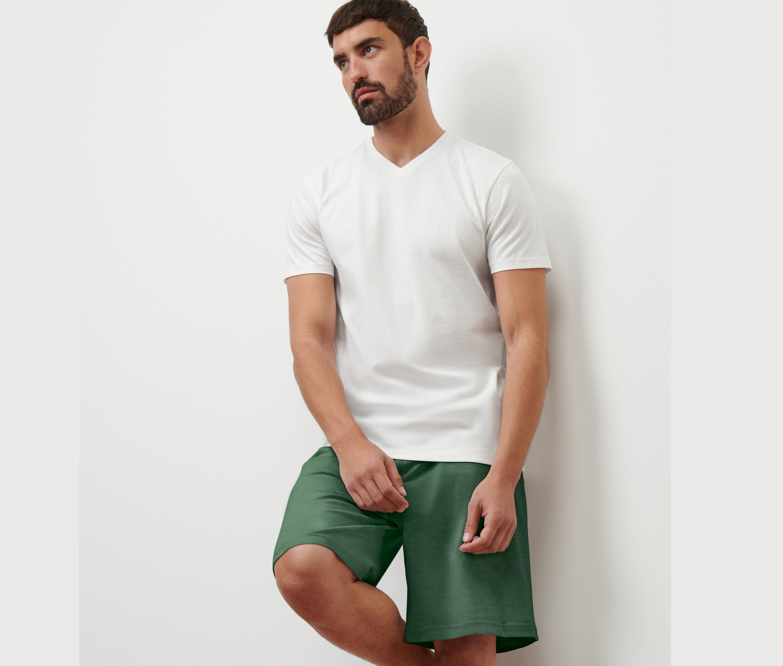Duvara yaslanmış bir adam beyaz V yaka tişört ve yeşil şort giyiyor. 2 Adet V Yaka Tişört'ten 1 tanesini giyiyor.