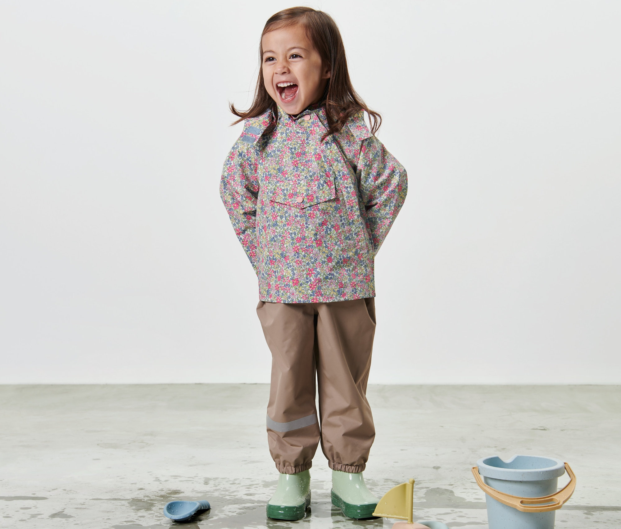 Gülümseyen kız çocuğu çiçek baskılı çocuk yağmurluğuyla yerde oyuncaklarla oynuyor.