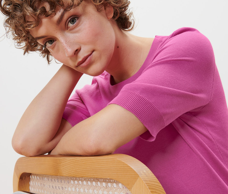 Kadın dirseğini sandalye sırtına yaslamış, pembe ince örgülü kazak giyiyor.