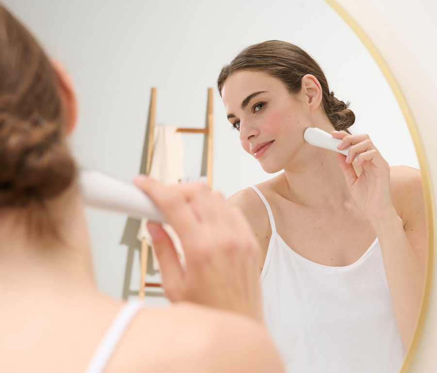 Kadın aynanın önünde elektrikli yüz epilatörü kullanıyor.