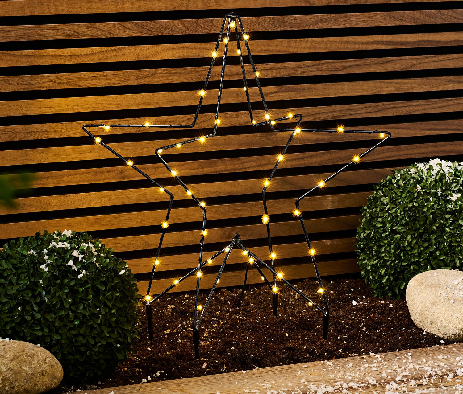 Metal LED dış mekan yıldızı yerde çalıların ve taşların yanında duruyor.