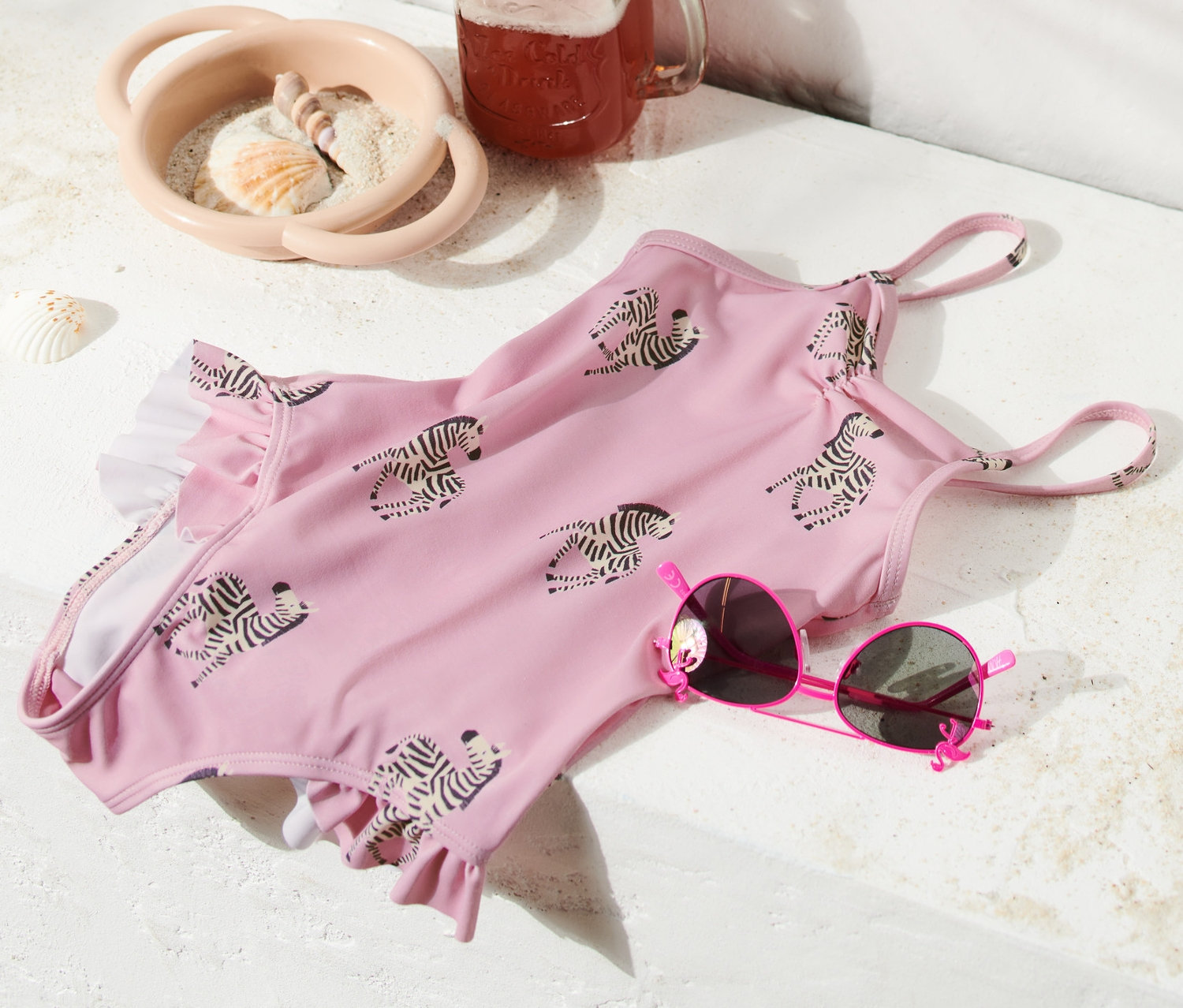 Zebra desenli, fırfırlı pembe bir mayo, pembe güneş gözlüğü, içecek olan bir bardak ve bir plaj çantası yanında duruyor.