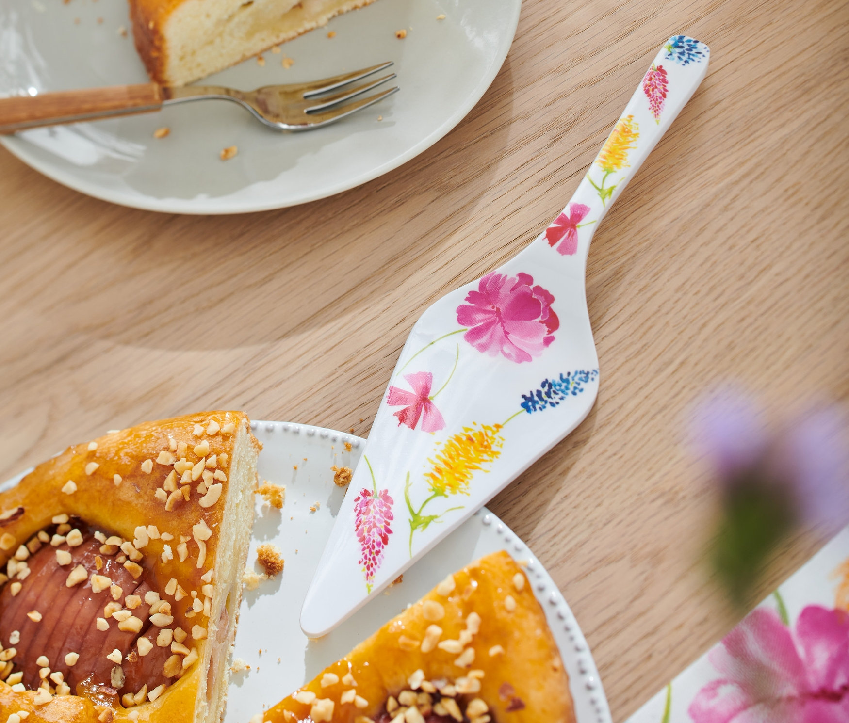 Bir masanın üzerinde bir dilim kekin bulunduğu bir tabak, bir çatal ve çiçek desenli bir pasta spatulası, yanında da bütün bir kek duruyor.