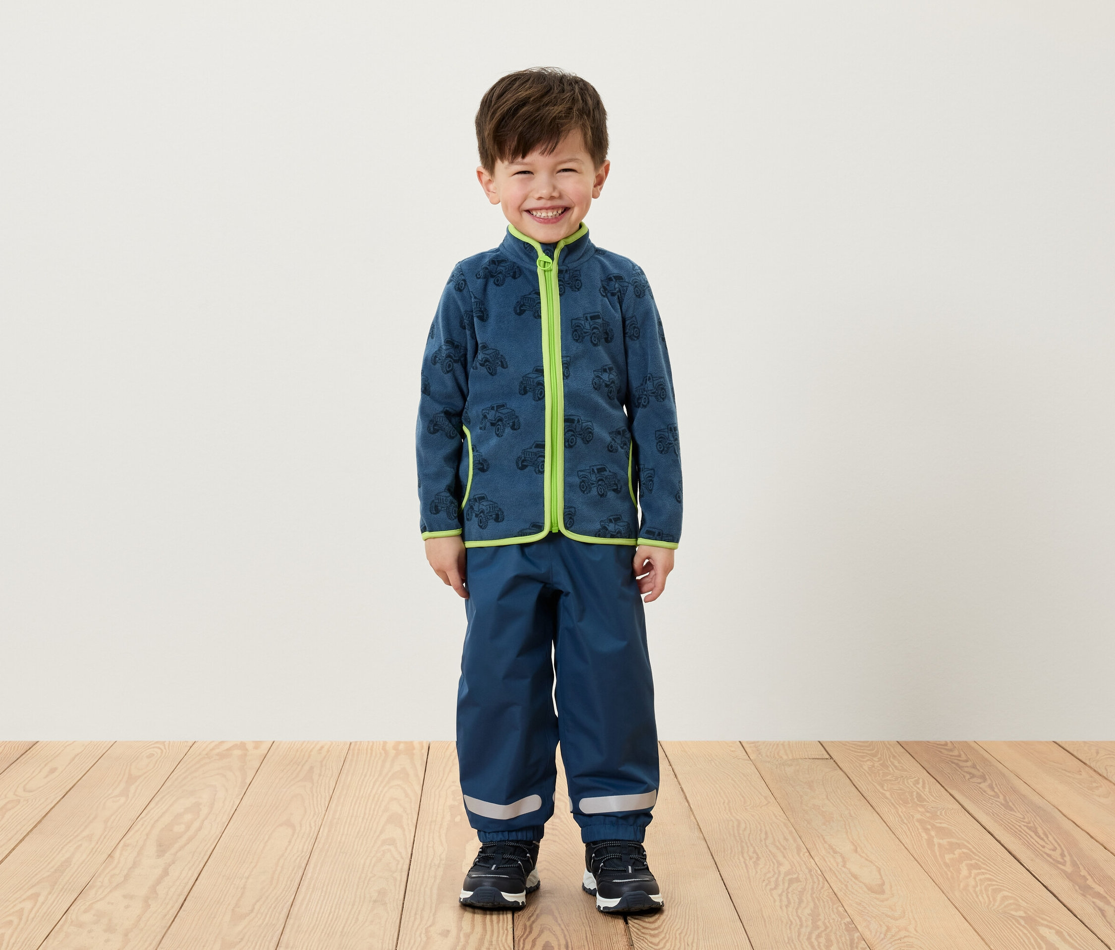 Bir çocuk ayakta duruyor, üzerinde yeşil detayları olan mavi bir polar ceket ve mavi pantolon var.