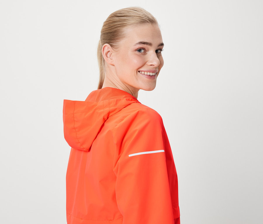 Bir kadın omzunun üzerinden gülümsüyor, kapüşonlu ve reflektörlü turuncu bir ceket giyiyor.