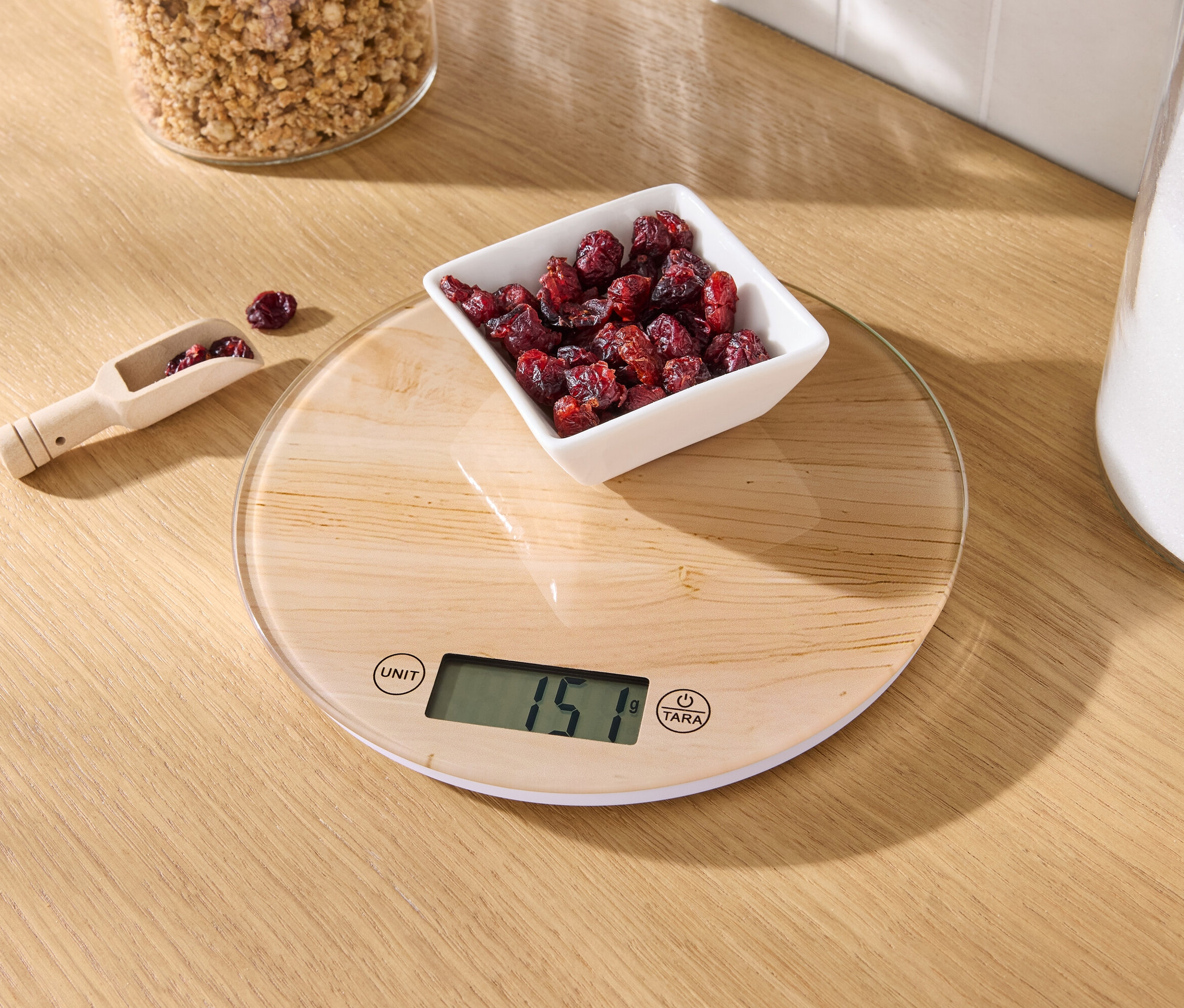 Ahşap bir masanın üzerinde, bir kase kuru kızılcık ve kızılcık bulunan ahşap bir ölçü kaşığı ile birlikte bir mutfak terazisi bulunmaktadır.