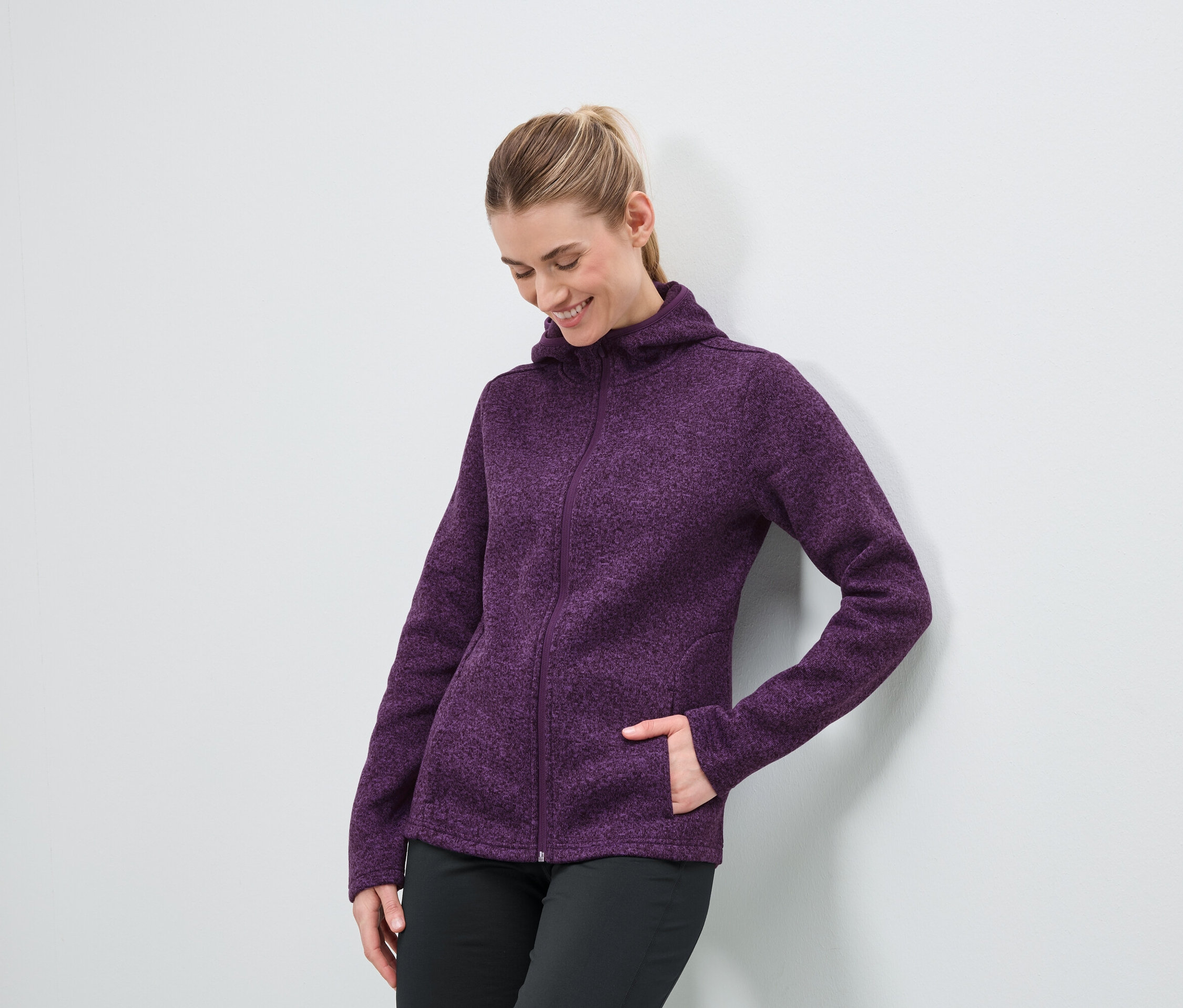 Mor renkli fermuarlı kapüşonlu ve siyah pantolonlu bir kadın beyaz bir duvarın önünde duruyor.