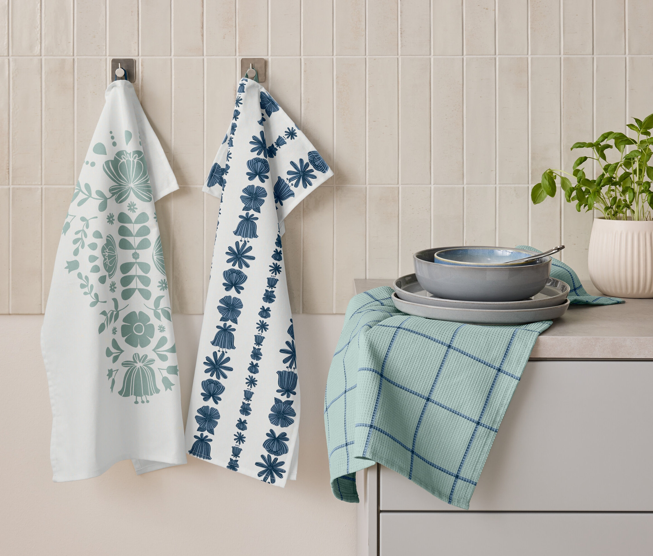 Çiçek desenli iki adet mutfak havlusu duvarda yan yana asılı duruyor. Altında, tabakların ve bir kaseğin üst üste konduğu, mavi çizgili katlanmış yeşil bir mutfak havlusu duruyor. Yanında ise bir saksı bitki bulunuyor.