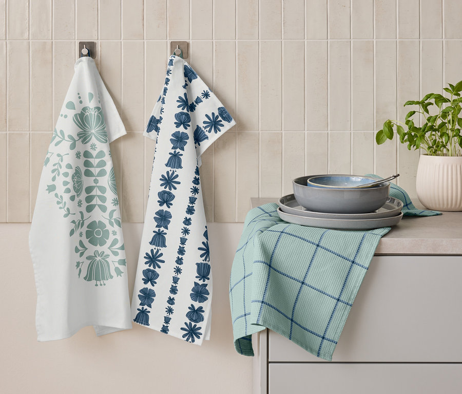 Çiçek desenli iki adet mutfak havlusu duvarda yan yana asılı duruyor. Altında, tabakların ve bir kaseğin üst üste konduğu, mavi çizgili katlanmış yeşil bir mutfak havlusu duruyor. Yanında ise bir saksı bitki bulunuyor.