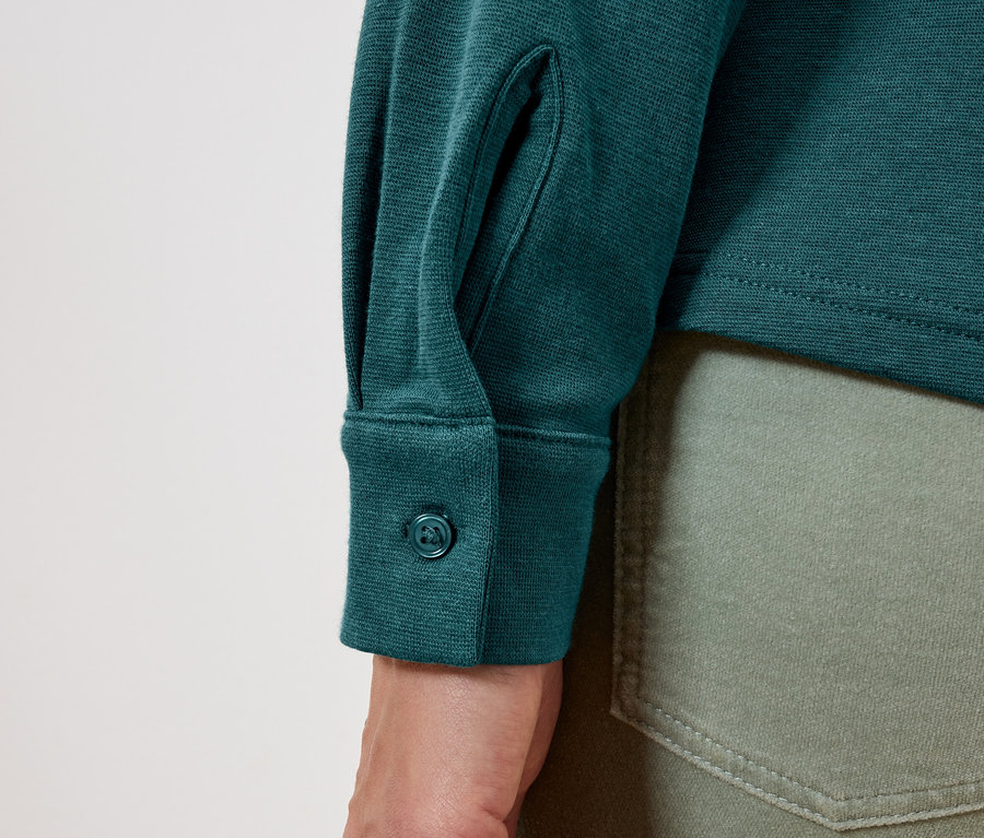 Manşetinde düğmesi olan uzun kollu yeşil bir gömlek ve zeytin yeşili kot pantolon detayı.