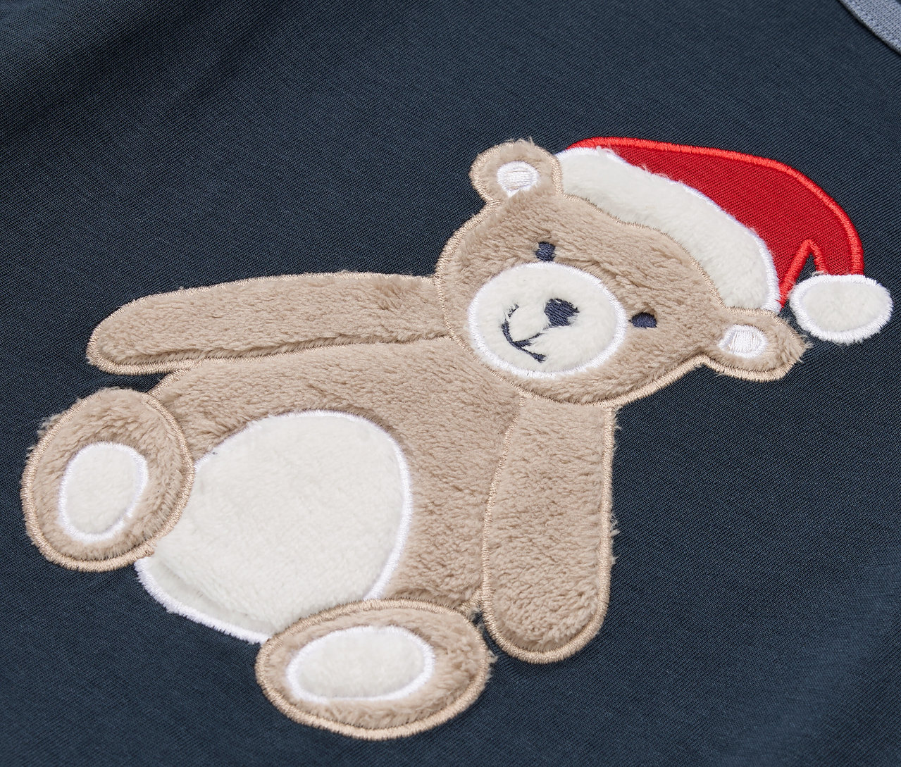 Koyu mavi bir kumaş üzerinde kırmızı Noel şapkalı kahverengi bir oyuncak ayı bulunmaktadır.