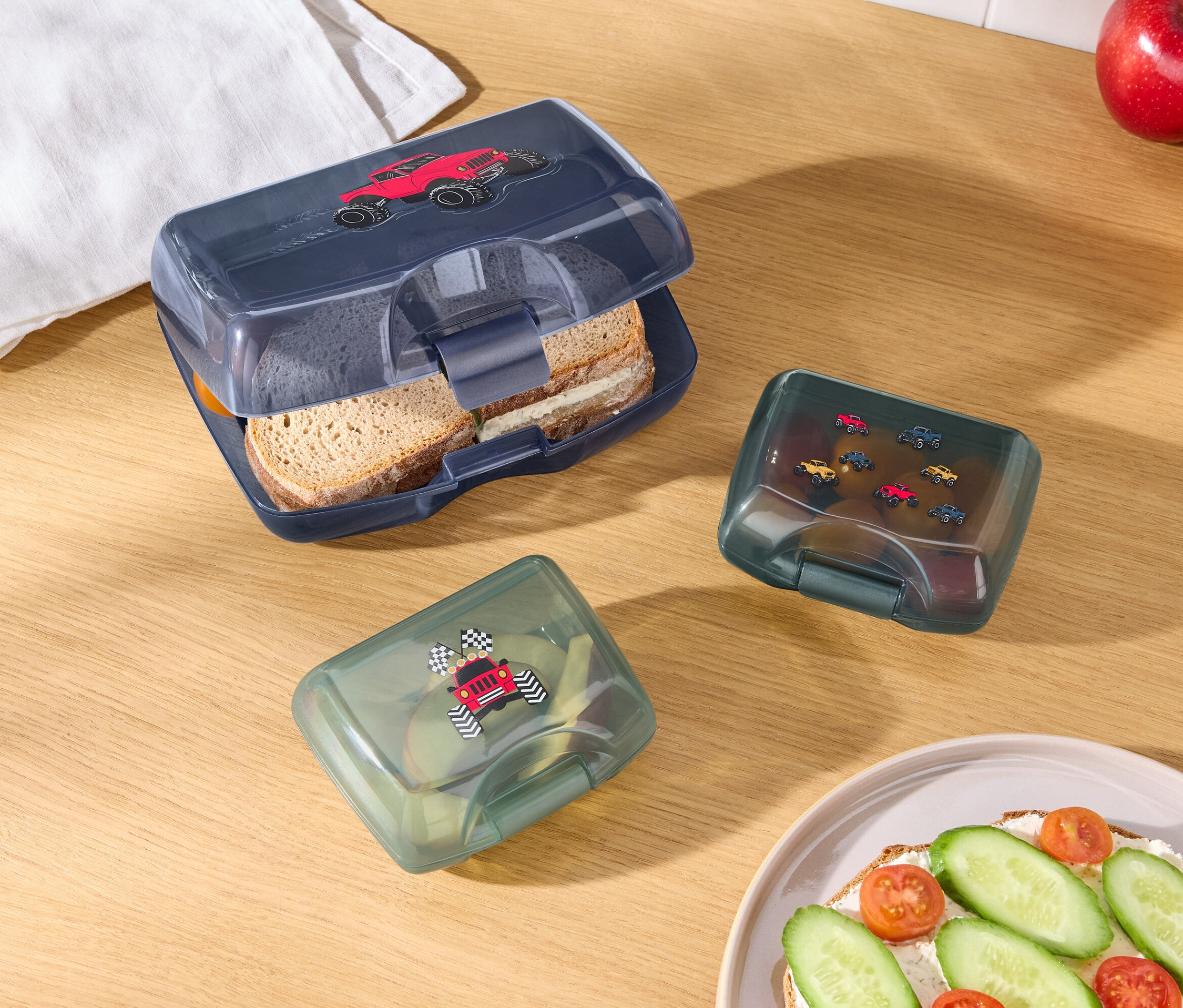 Ahşap bir masa üzerinde kırmızı araba resimli açık mavi bir beslenme kutusu, araba resimli iki adet yeşil beslenme kutusu, bir elma, bir portakal, beyaz bir bez ve sandviçli bir tabak bulunmaktadır.