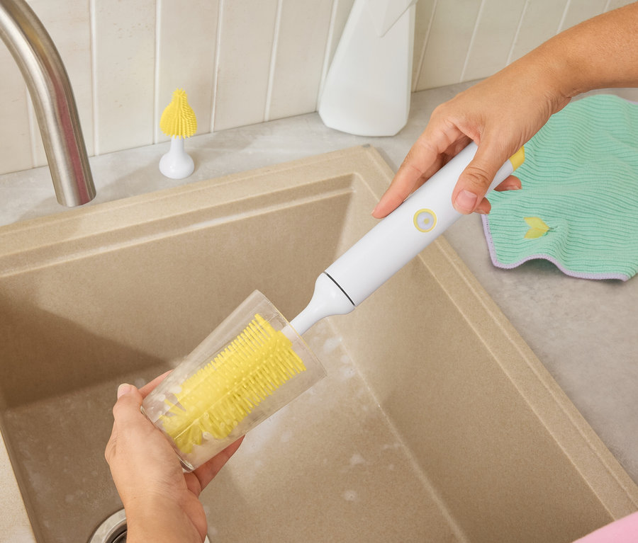 Bir kişi bej bir lavaboda sarı bir fırça ile bir bardağı temizliyor. Arka planda beyaz bir şişe ve yeşil bir bez var.