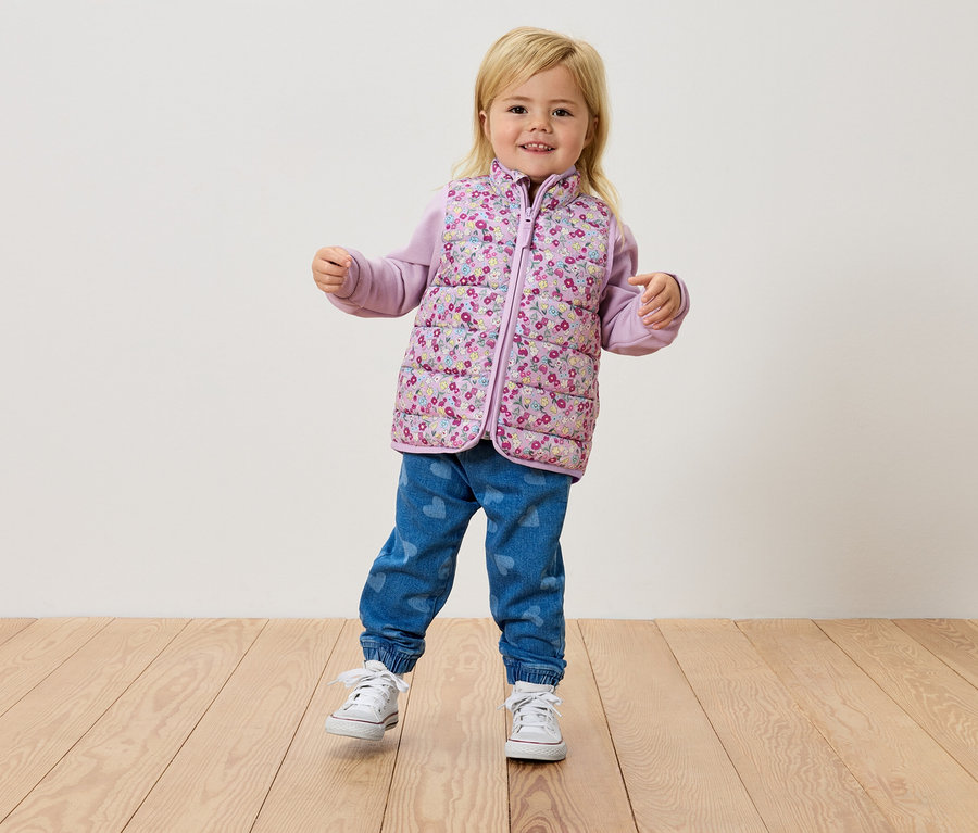 Küçük sarışın bir kız, tahta bir zeminde duruyor, çiçekli bir yelek, mor bir üst ve kot pantolon giyiyor.
