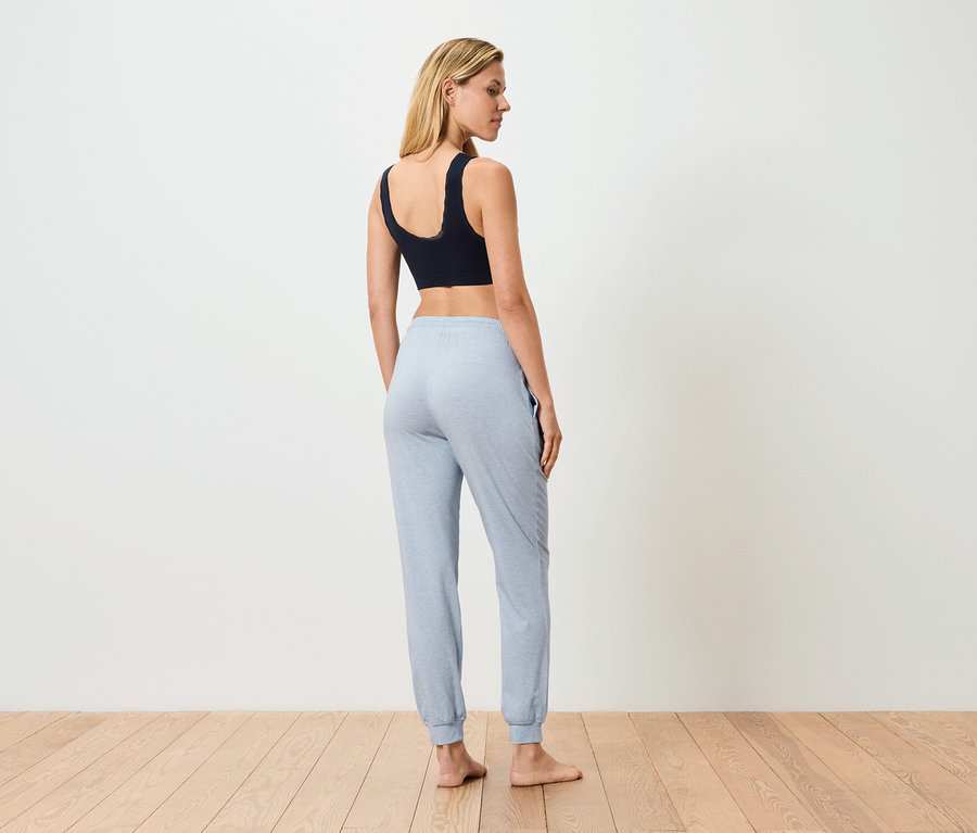 Kadın sırtı dönük duruyor, siyah bir spor sütyeni ve açık mavi eşofman altı giyiyor.