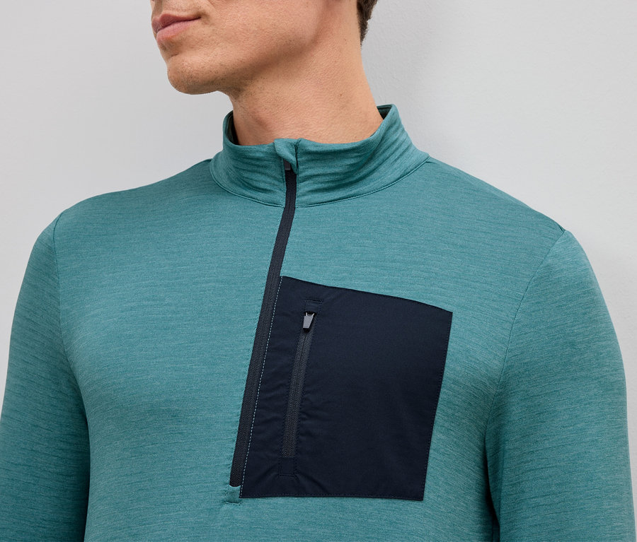 Yakın çekimde, turkuaz uzun kollu, kısa fermuarlı ve sol tarafında tek yama fermuarlı cebi olan bir adam.