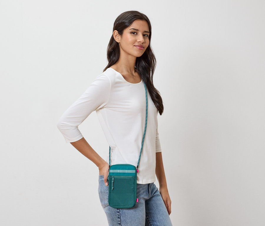Koyu kahverengi saçlı bir kadın, turkuaz askılı yeşil bir çapraz çanta, beyaz uzun kollu bir gömlek ve kot pantolon giyiyor.
