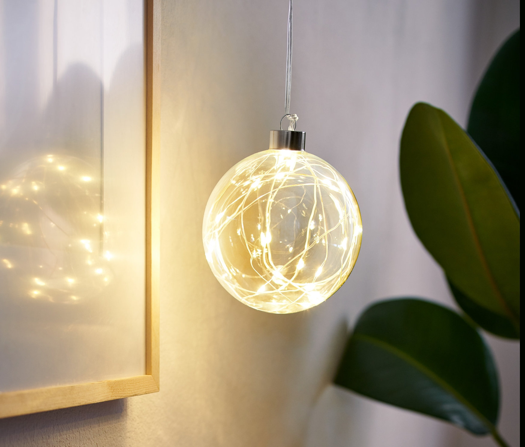 Bir LED Cam Küre odada asılı duruyor, yanında çerçeveli bir resim ve içinde otlar olan bir vazo var.