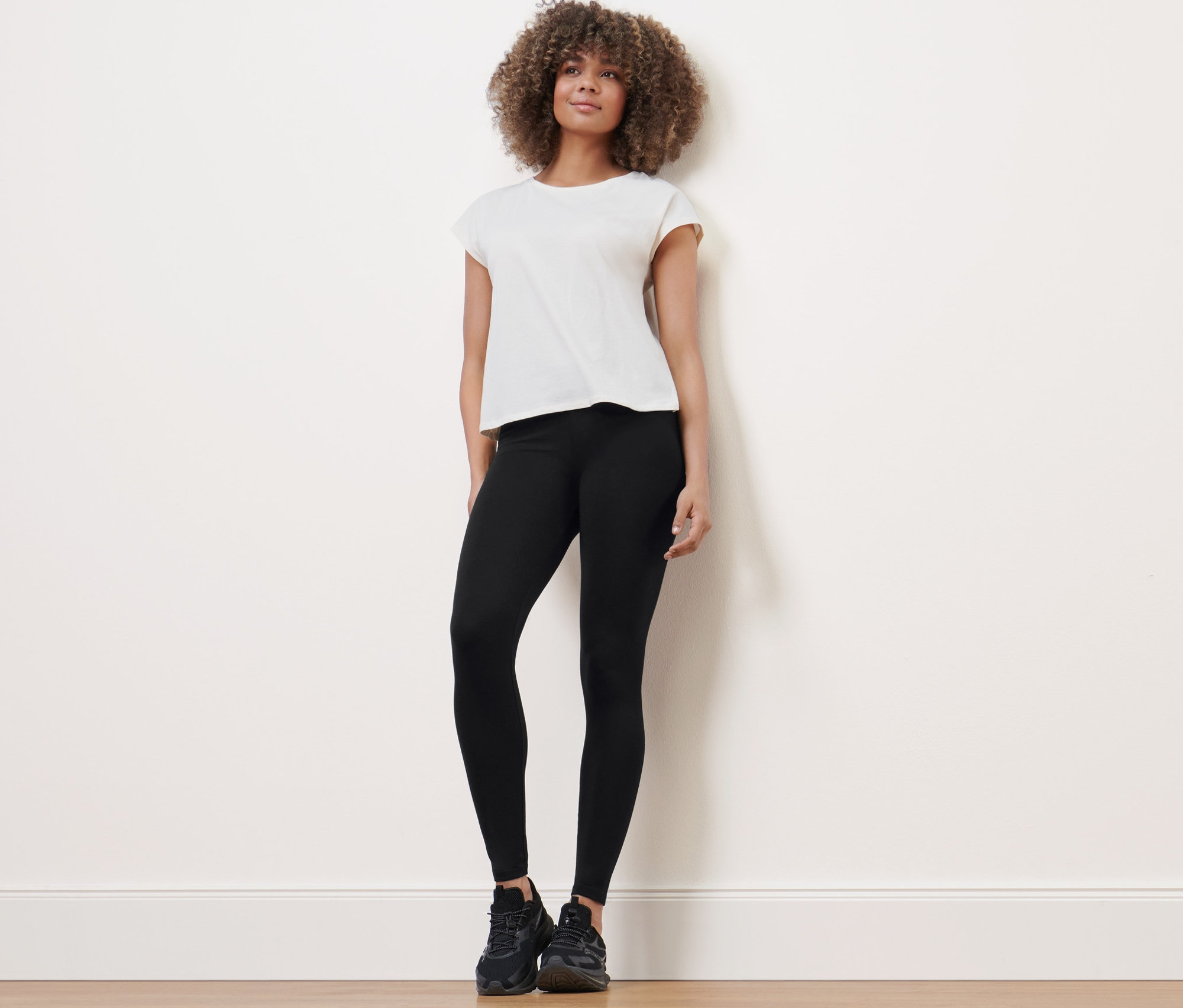 Kadın ayakta duruyor, spor taytı, tişört ve spor ayakkabı giyiyor.